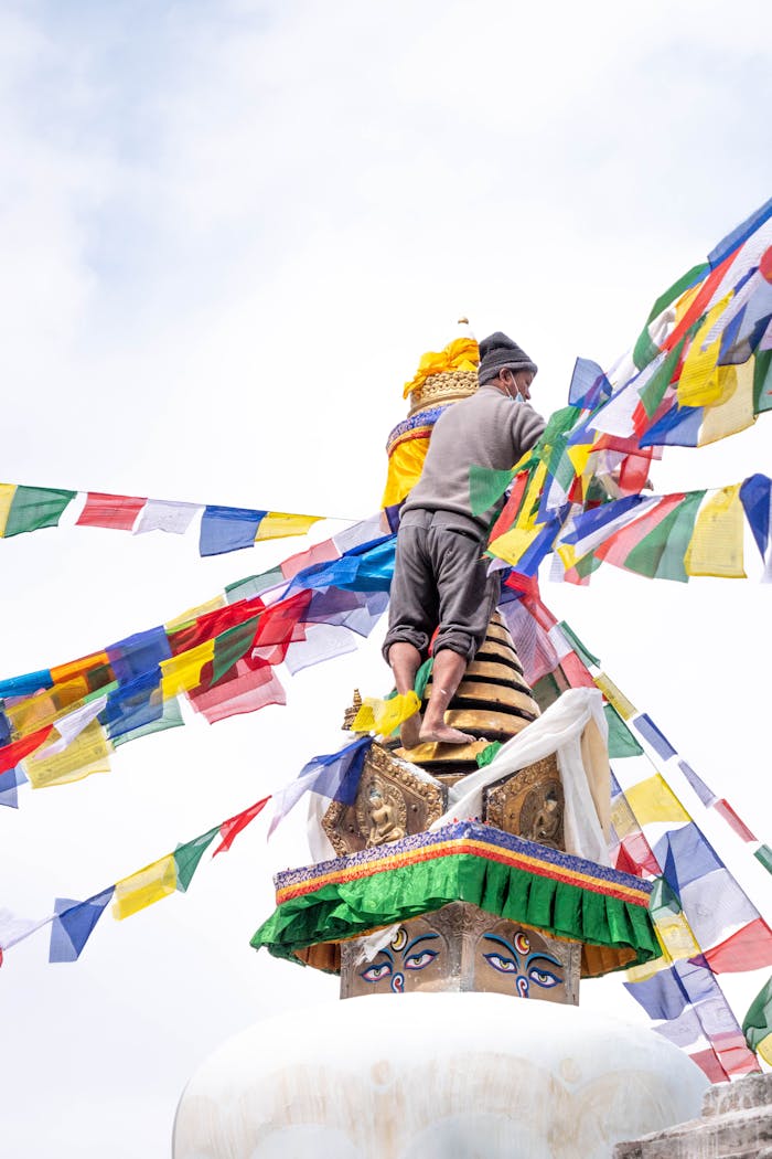 A man adjusting colorful prayer flags on a Tibetan Buddhist stupa under a clear sky.